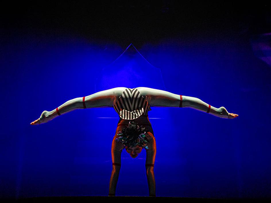 Cirque du Soleil contortionist balances in a deep backbend pose against a dark blue stage background.