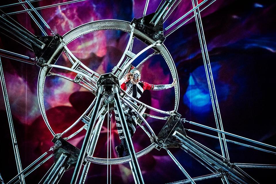 "Cirque du Soleil acrobat performs inside a large rotating metal wheel against a vivid, abstract digital background.