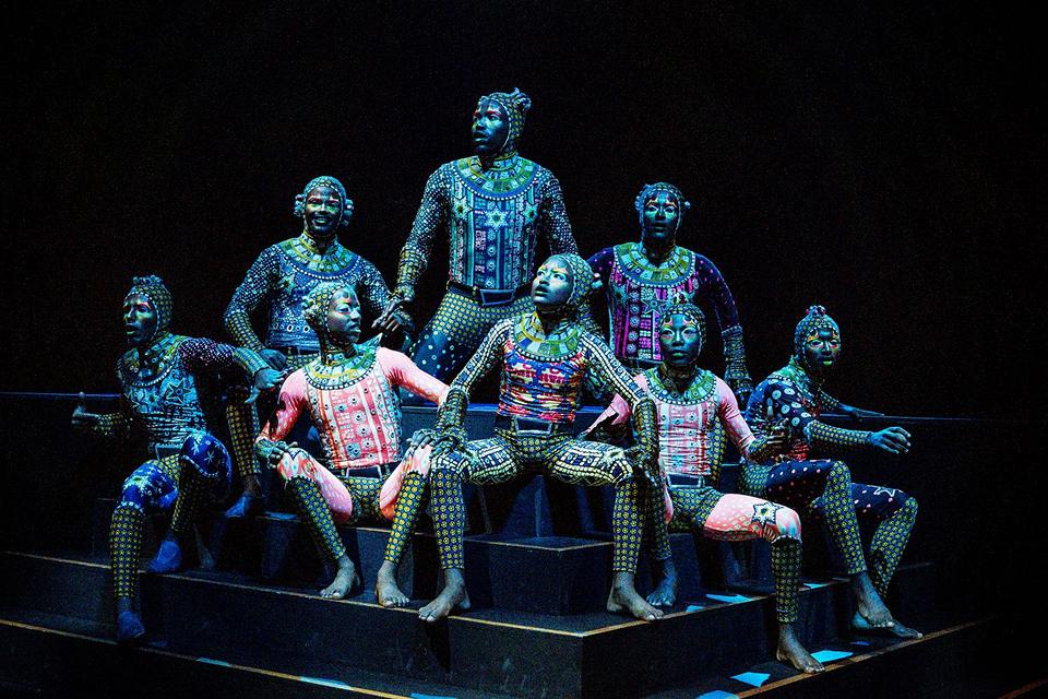 Cirque du Soleil ensemble of performers in detailed, body-painted costumes pose together on tiered platforms under dramatic lighting.