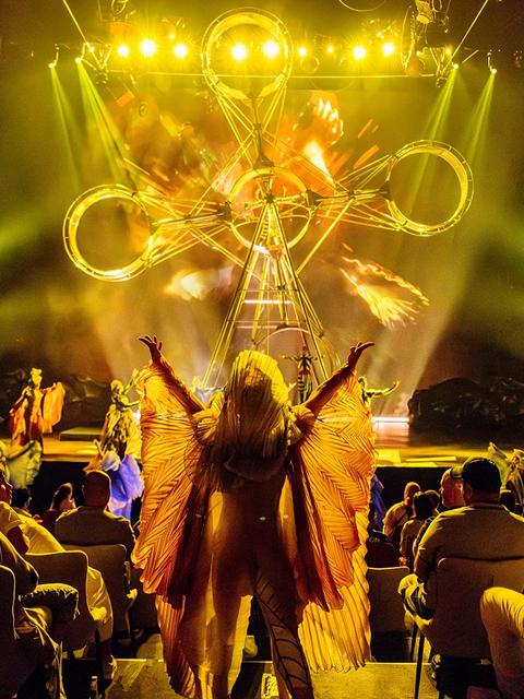 Cirque du Soleil performer with illuminated wings stands in front of a spectacular stage set with fire effects and golden lighting.