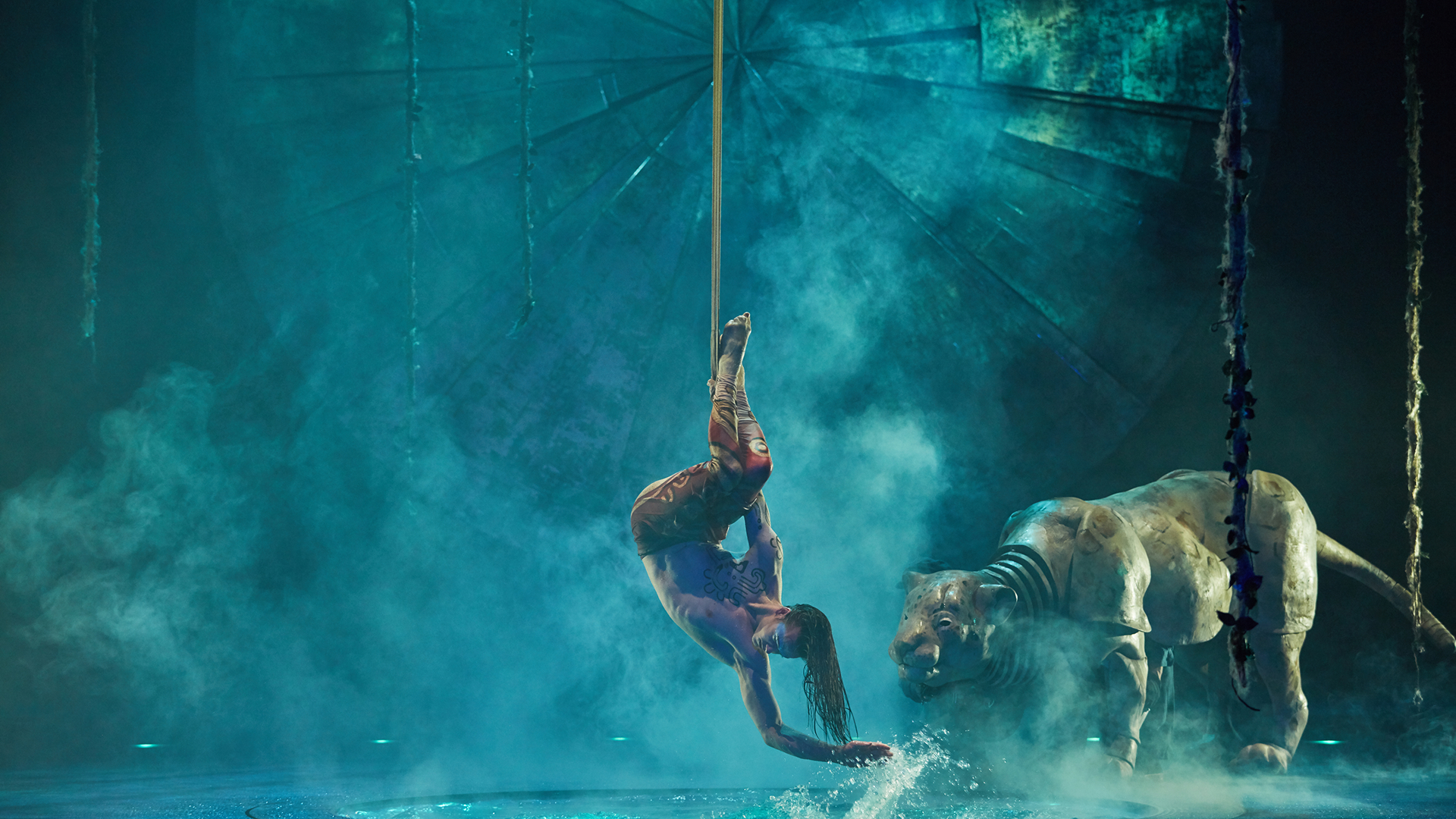 A Cirque du Soleil aerial performer hangs upside down from a rope above water, performing beside a life-sized tiger sculpture in a dramatic, mist-filled stage setting.